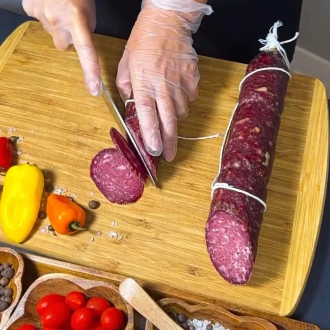 Dry-cured beef sausage being sliced on a wooden board with vegetables and spices