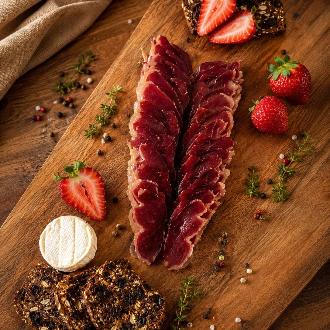 Thinly sliced dry-cured duck breast served on a rustic wooden board with artisan seed bread, creamy cheese, and fresh strawberries.