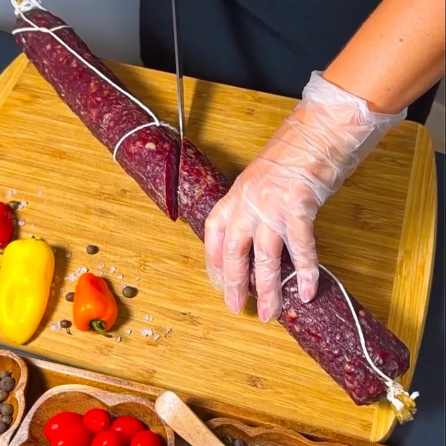Dry-cured beef sausage being sliced on a wooden board with vegetables and spices
