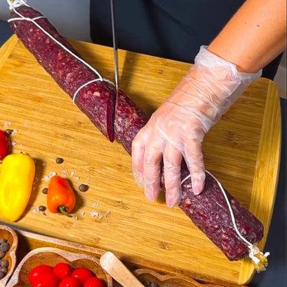 Dry-cured beef sausage being sliced on a wooden board with vegetables and spices