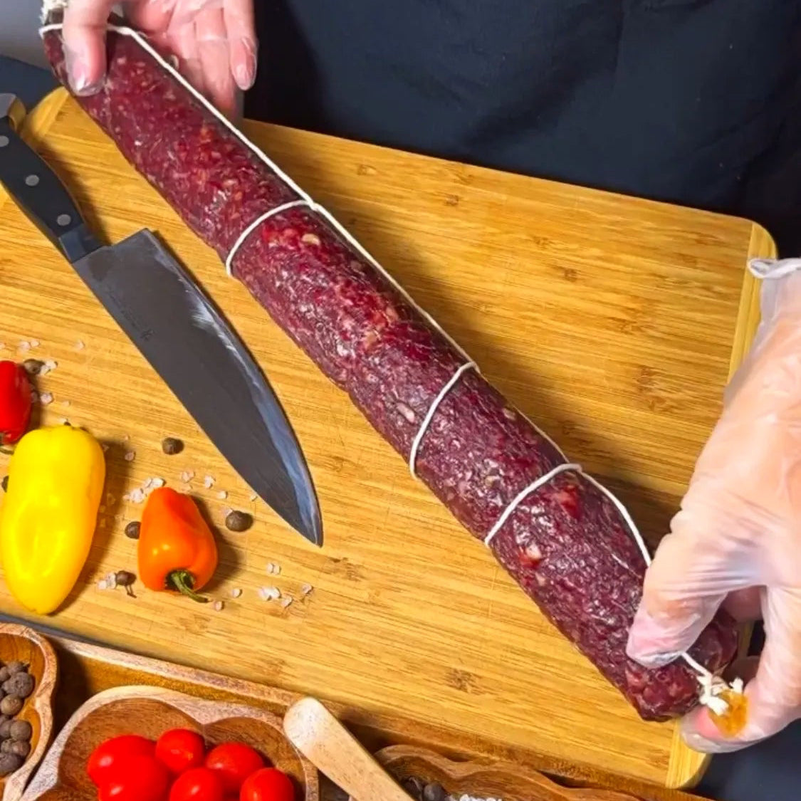 Whole dry-cured beef sausage on a wooden board with knife, vegetables, and spices